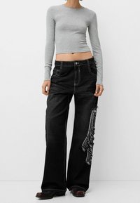 Mustat leveälahkeiset farkut valkoisillaommelluksilla ja monimutkaisella sivustakirjailulla. Yhdistetty tiukkaan harmaaseen pitkähihaiseen paitaaan.