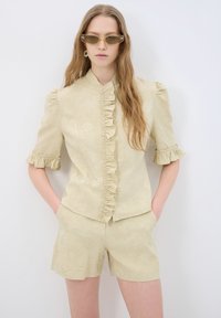 Top beige chiaro con dettagli arricciati sul davanti e maniche a sbuffo corte, abbinato a pantaloni corti coordinati. Il tessuto presenta un motivo floreale sottile. Vestibilità aderente.