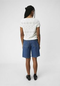 Weißes kurzärmliges Oberteil mit Lochmuster, kombiniert mit blauen Denim-Shorts. Die Rückansicht zeigt die Textur des Shirts sowie die Länge und Taschen der Shorts.