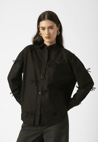 Black button-up shirt with bow accents along sleeves and front. Made of a textured fabric, featuring a classic collar and relaxed fit.