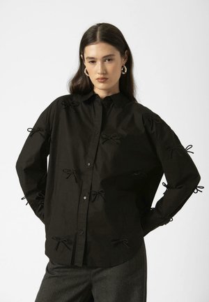 Black button-up shirt with bow accents along sleeves and front. Made of a textured fabric, featuring a classic collar and relaxed fit.