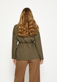 Olivovo zelená bunda s opaskom, dlhé rukávy a priliehavý strih, v kombinácii s hnedými nohavicami s širokými nohavicami. Hladká textúra, minimalistický dizajn.