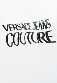 Μαύρο στυλιζαρισμένο κείμενο που αναγράφει "Versace Jeans Couture" σε λευκό φόντο με ελαφρώς φθαρμένη υφή.