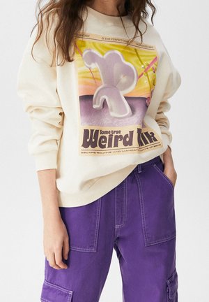 Femme portant un sweat-shirt crème avec un graphisme abstrait et un texte "some true weird life", associé à un pantalon violet avec de grandes poches.
