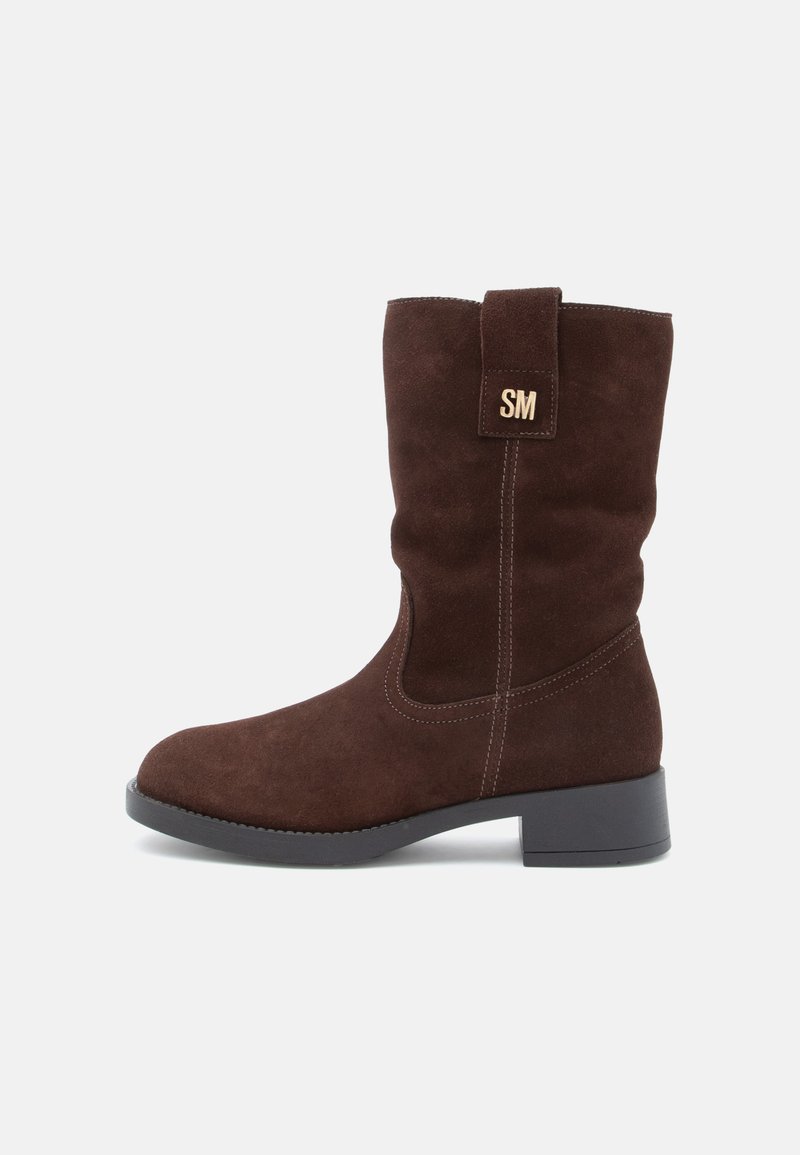 Bottes hautes en daim marron avec un talon bloc, présentant un design cousu et une boucle décorative avec les lettres "SM" en or.