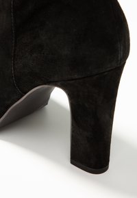 Botte en daim noir avec un talon épais sur une surface blanche, montrant des textures et des détails de couture.