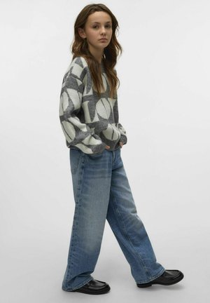 Grau-weißer, gemusterter Pullover mit voluminösen Ärmeln, kombiniert mit lockeren blauen Jeans und schwarzen Loafers. Heller Hintergrund.