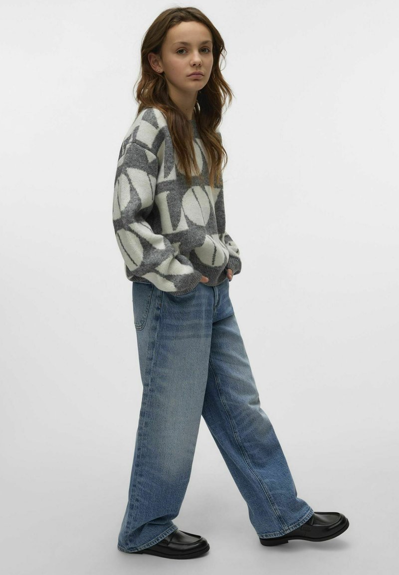 Pull gris et blanc à motifs avec des manches volumineuses, associé à un jean bleu ample et des mocassins noirs. Fond clair.