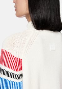 Knitted sweater in cream with color-blocked striped accents in red, blue, and black. Features a high collar and a small logo patch at the back.