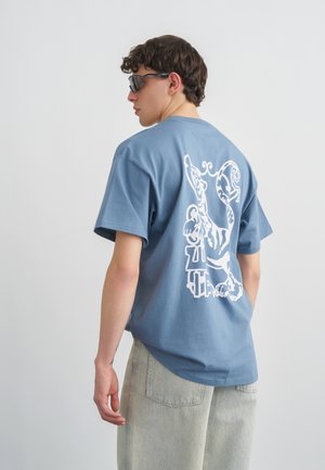 Młody mężczyzna noszący okulary przeciwsłoneczne, niebieską koszulkę z białym abstrakcyjnym wzorem na plecach oraz jasne jeansy, zwrócony tyłem do kamery.