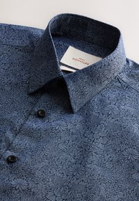 Tumesinine särk tekstuurse lillemustriga, nööbitav krae ja mustad nupud. Sildil on kiri „NEXT SIGNATURE.”