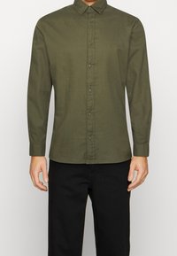 Długi rękaw oliwkowej zielonej koszuli z zapięciem na guziki z przodu, klasycznym kołnierzem, miękką fakturą i dopasowanym krojem. W zestawie z czarnymi spodniami.