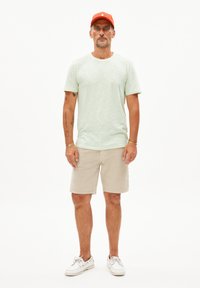 Hellgrünes, strukturiertes T-Shirt, beiger Shorts und weiße Schuhe. Trägt eine orange Mütze, mit sichtbaren Tattoos auf den Armen. Steht vor einem weißen Hintergrund.