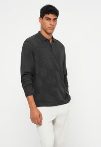 Langärmliges schwarzes Poloshirt mit subtilen strukturierten Mustern, ausgestattet mit einem Kragen und einer Knopfleiste, kombiniert mit hellen Hosen.