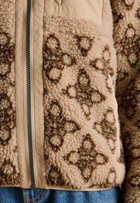 Beige and brown patterned fleece jacket with a visible vertical zipper, worn over blue denim jeans.