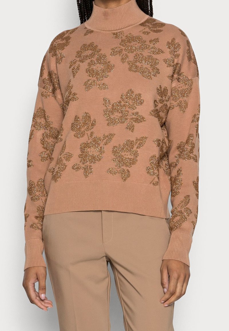 Pull en tricot rose avec un col montant, orné de motifs floraux pailletés dorés et des poignets et ourlet côtelés. Associé à un pantalon beige.
