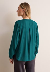 Blusa lunga color turchese a maniche lunghe con una vestibilità rilassata, dotata di una piega sulla schiena e una texture morbida. L'orlo è leggermente curvato, complementando dei pantaloni larghi.