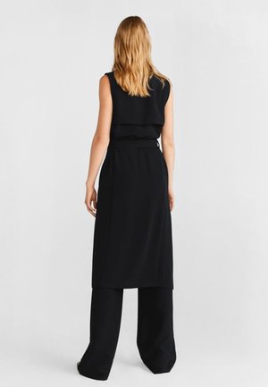 Femme aux cheveux blonds portant une robe noire sans manches par-dessus un large pantalon noir, debout de dos sur un fond uni.