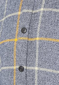 Blauw en grijs geruit stof met gele en witte lijnen, met een soepele textuur en een zwart knoopdetail op de overslag.