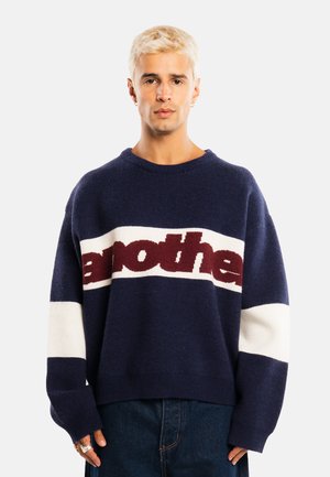 Jeune personne aux cheveux blonds courts porte un pull bleu marine avec des rayures horizontales blanches et bordeaux ainsi qu'un texte, associé à un jean foncé.
