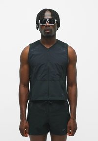 Schwarze sportliche Weste mit Reißverschluss, aus leichtem Stoff und mit kurzen Ärmeln, kombiniert mit schwarzen Shorts. Dazu werden schwarze Sonnenbrillen getragen.