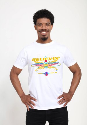 Smiling man with curly hair wears white t-shirt featuring colorful winged scarab beetle and text "Don't Stop Believin' Journey."