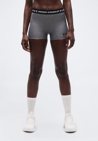 Αθλητικό άτομο που φορά γκρι σορτς Under Armour, στέκεται με λευκές κάλτσες και λευκά αθλητικά παπούτσια μπροστά σε απλό φόντο.