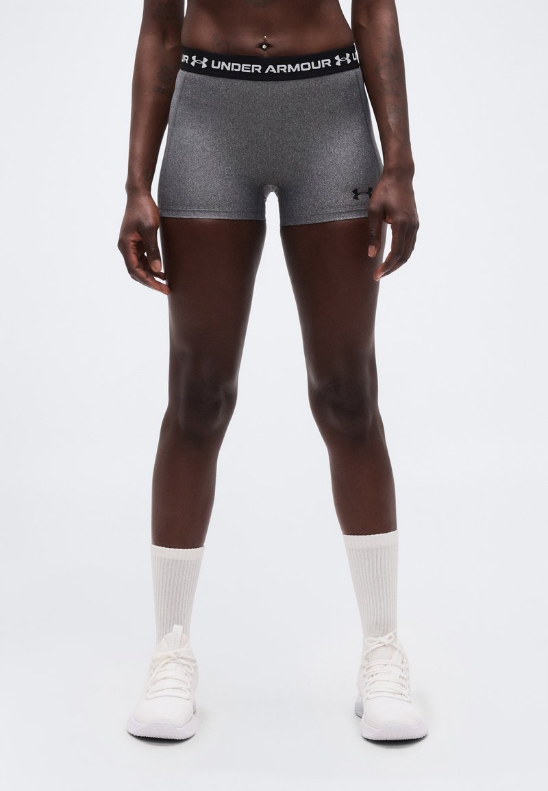 Αθλητικό άτομο που φορά γκρι σορτς Under Armour, στέκεται με λευκές κάλτσες και λευκά αθλητικά παπούτσια μπροστά σε απλό φόντο.