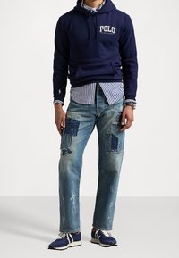 Námořnická bavlněná mikina s logem "Polo", vrstvená přes modro-bíle pruhovanou košili. Nosí se s roztrhanými džíny s detailními nášivkami.