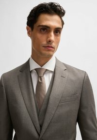 Costume trois pièces gris avec un revers cranté, gilet assorti et chemise blanche. Accentué par une cravate à pois marron clair. Texture de tissu lisse.