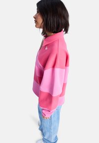 Fleece-genser med krage, med horisontale striper i varierende nyanser av rosa. Inkluderer en liten logo på brystet.