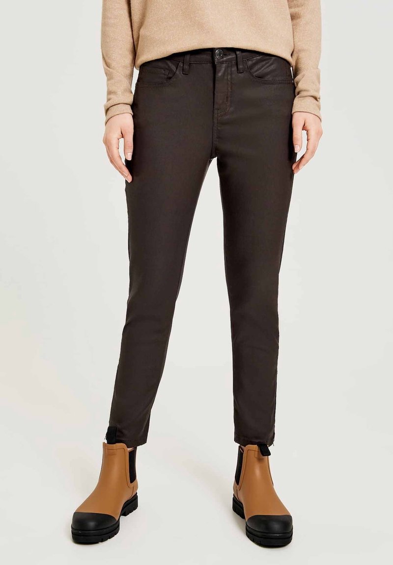 Personne portant un pantalon slim-fit marron foncé, un haut à manches longues beige et des bottines beige avec bouts noirs, se tenant sur un fond neutre.