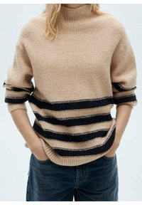 Pull en rib d'un beige avec un col montant, présentant des rayures horizontales bleu marine et dorées. Texture douce avec des manches retroussées et une coupe décontractée.