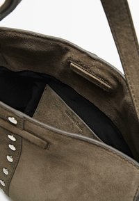 Braune Wildleder-Handtasche mit silbernen Nieten, geöffnet, um das schwarze Stofffutter und das Markenetikett in der Innentasche zu zeigen.