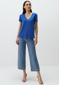 Niebieska koszulka z dekoltem w serek, wykonana z miękkiego materiału, w połączeniu z szerokimi jasnoniebieskimi dżinsami. Model stoi na neutralnym, jasnym tle.