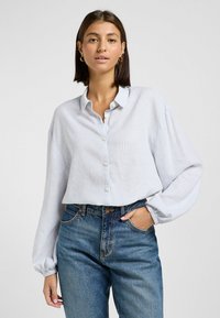 Camicia a righe azzurro chiaro e bianca con colletto, vestibilità ampia e polsini elasticizzati, abbinata a jeans blu a vita alta.