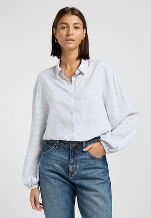 Camisa de botón a rayas en azul claro y blanco, con cuello, corte holgado y puños elásticos, combinada con unos vaqueros azules de cintura alta.