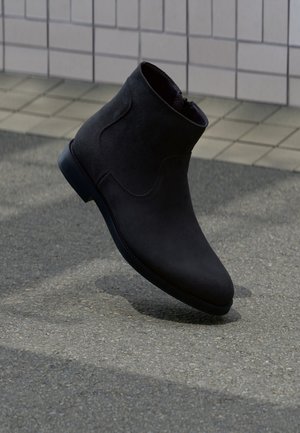 Stivaletto in camoscio nero che fluttua sopra l'asfalto vicino a una parete rivestita di piastrelle quadrate color grigio chiaro e beige.