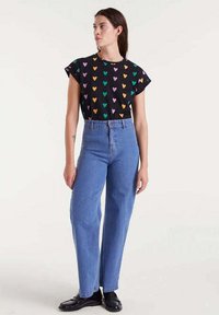 Mujer de pie, vestida con una camiseta negra de manga corta con corazones multicolores, unos vaqueros azules de tiro alto y unos mocasines negros, frente a un fondo liso.