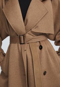 Trench-coat en laine marron avec large col, boutons noirs et ceinture avec boucle noire rectangulaire sur un haut noir.