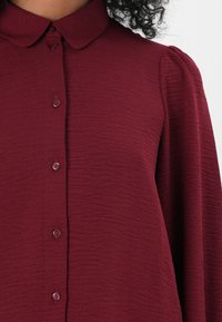 Bluză burgundy cu mânecă lungă, guler, țesătură texturată, nasturi pe față și mâneci ușor bufante.
