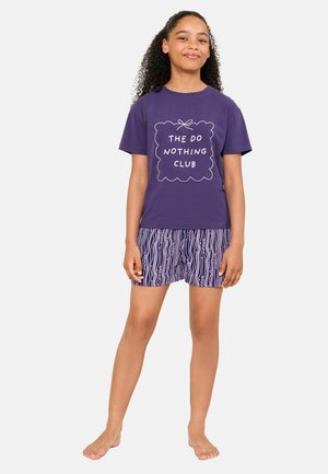 Pige med krøllet hår står barfodet iført en lilla T-shirt med teksten "The Do Nothing Club" og stribede shorts mod en ensfarvet baggrund.