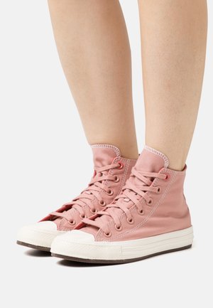Baskets montantes en toile rose avec un bout en caoutchouc blanc et une semelle blanche, présentant des œillets et des lacets rouges. Texture lisse, branding minimal.