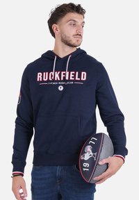 Un sweat à capuche bleu marine en coton, avec le logo "Ruckfield" en rouge et blanc, doté d'une poche kangourou et de cordons de serrage. Accompagné d'un ballon de rugby.