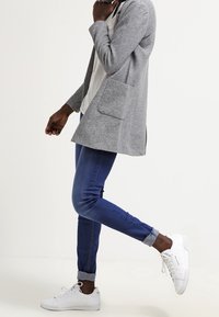 Grå stickad cardigan med två framfickor, bärs över en vit topp. Matchas med blå jeans och vita sneakers. Slät textur och modern passform.