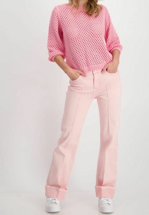 Femme portant un pull rose en maille ajourée, un jean large rose clair avec des revers, des baskets blanches, debout les mains dans les poches.
