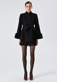 Cappotto nero con cintura in vita, maniche lunghe e polsini in pelliccia sintetica. Tessuto strutturato, design aderente, abbinato a pantaloni shorts neri e tacchi lucidi color bordeaux.