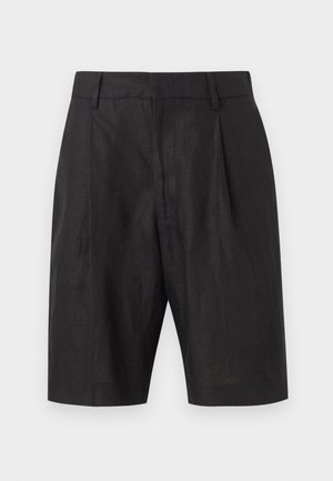 Homme aux cheveux courts et bouclés portant une chemise à manches courtes à motif orange rouille, un short noir au genou et des sandales noires faciles à enfiler.