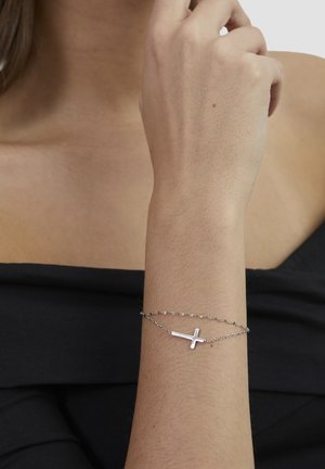Woman wearing a delicate silver chain bracelet with a horizontal cross charm on her wrist, dressed in a black off-shoulder top.
