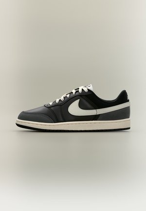 Niski czarno-szary sneaker Nike z białym znakiem, białymi sznurówkami i beżową podeszwą na neutralnym tle.